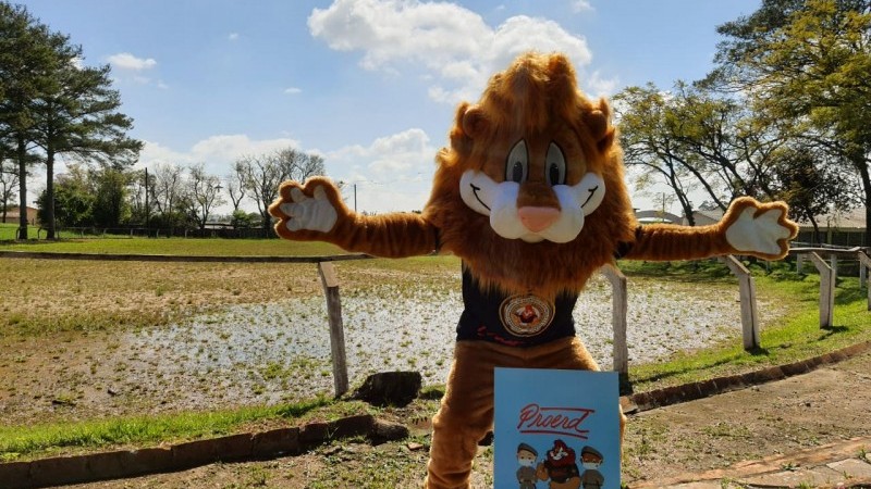Em primeiro plano o mascote do Leão do PROERD está posando de braços abertos num campo ao ar livre.