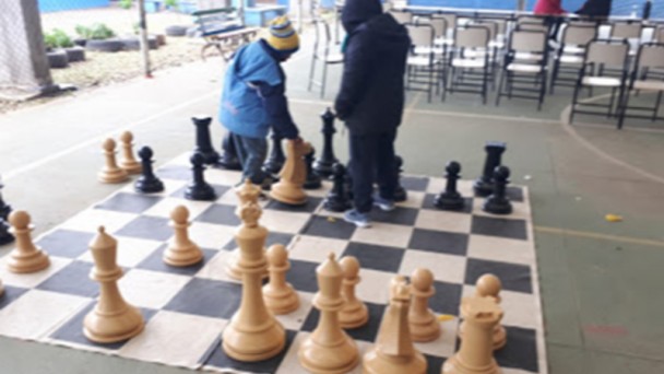 No pátio de uma escola, crianças movimento grande peças de xadrez sobre um tabuleiro quadriculado preto e branco estendido no chão de uma quadra de esportes.