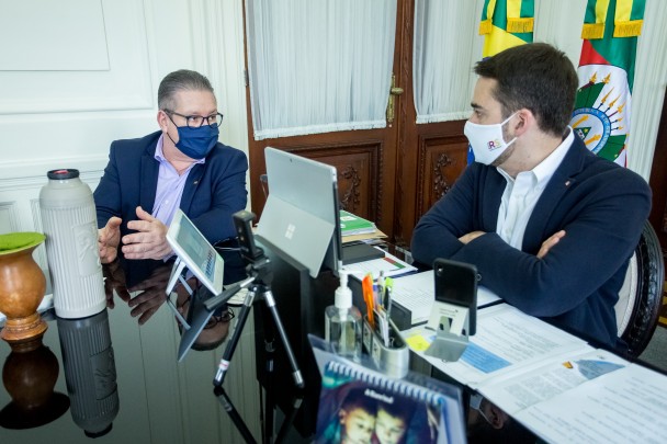 Sentados diante de uma mesa de trabalho com notebook, celular, tablet, cuia e térmica de chimarrão e documentos, o governador e o vice-governador conversam. Ambos estão de máscara contra a Covid-19. Ao fundo, bandeira do RS.