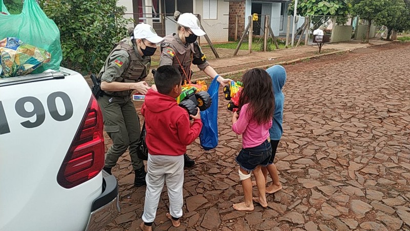 Duas policiais militares entregam presentes para três crianças, Todos estão em uma rua de paralelepipedos e no fundo da image é possível ver algumas casas