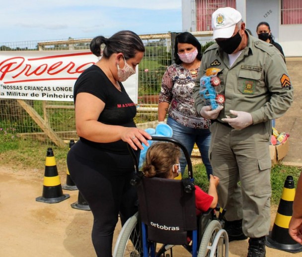 Uma criança cadeirante, de costas, recebe um brinquedo das mãos de um policial militar