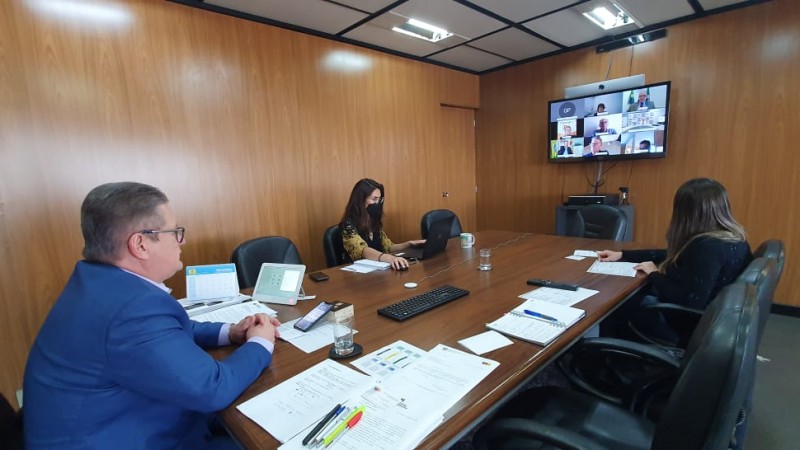 Vice-governador Ranolfo Vieira Júnior, sentado à ponta de uma grande mesa de reuniões em seu gabinete, observa ao fundo TV com imagem da videoconferência sobre a Operação RS Verão Total 2021.