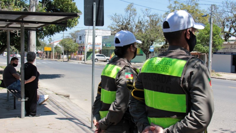 Dois policiais militares em pé, parados ao lado de um ponto de ônibus, observam o movimento na rua à frente.