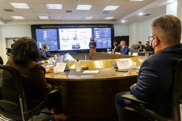 No fundo da imagem, de pé, a diretora geral do instituto geral de perícias a frente de um telão com dados, discursa para as pessoas em torno de uma grande mesa de reuniões