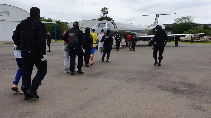 No pátio do hangar do Batalhão de Aviação da Brigada Militar, fila de presos escoltados por polícias federais durante embarque em avião da Polícia Federal que aparece ao fundo.