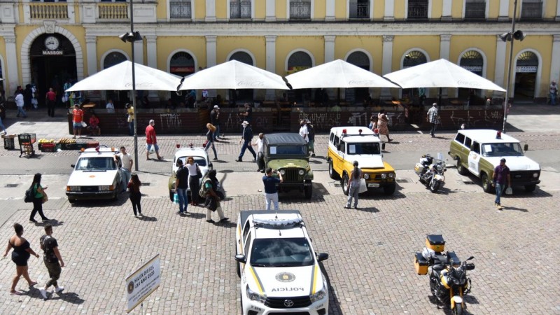 A imagem do alto mostra cinco viaturas antigas da Brigada Militar e uma nova viatura da Brigada Militar, pessoas caminham entre os carros estacionados