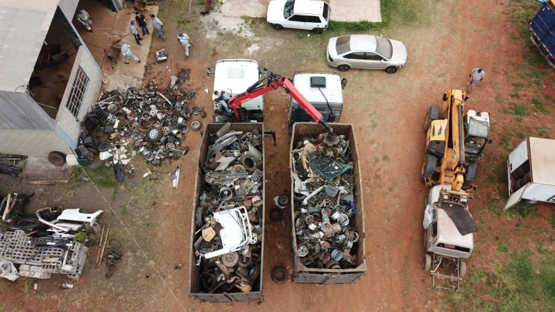 Visto do alto, duas caçambas de caminhão cheias de restos de veiculos, estacionados lado a lado em um chão de terra