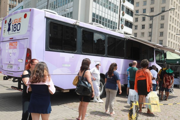 Fila de pessoas em frente a um ônibus lilás.