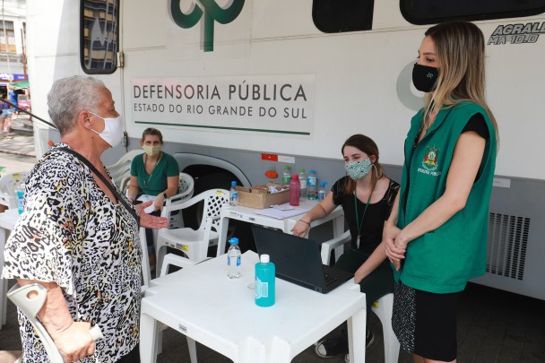 Em frente a um micro-ônibus da Defensoria Pública, uma servidora do órgão, de colete verde, conversa com uma idosa, de camisa com estampa tigrada, apoiada em uma muleta. 