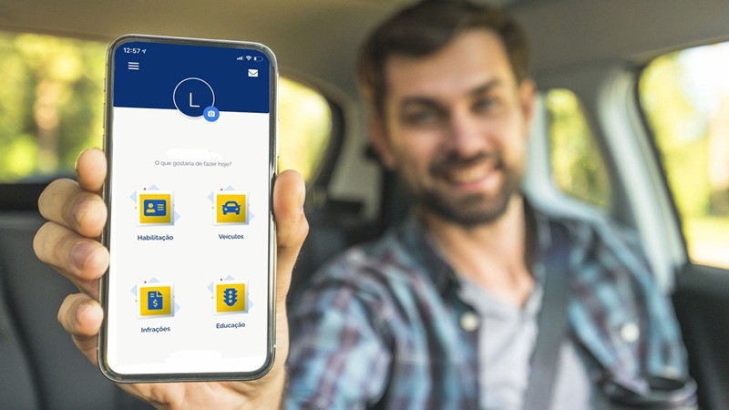 Homem sentado dentro de um carro, vestindo camiseta cinza e camisa xadrez por cima, usando o cinto de segurança, segura um celular com a mão direita. Na tela, a página inicial do app Carteira Digital de Trânsito.