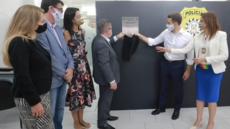 O governador Eduardo Leite, o vice-governador e secretário da Segurança Pública, Ranolfo Vieira Júnior, e a Chefe de Polícia, Nadine Anflor, estão juntos segurando um tecido inaugurando a placa da DPCI.
