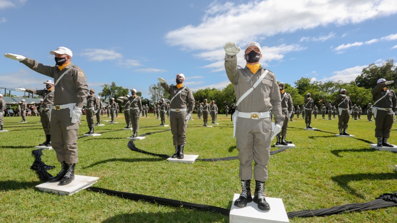 Policiais militares fardados em fila com o braço direito estendido à frente em um campo de futebol.