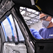 Na fábrica da Hich-Tech blindagens, em SP, visão a partir do interior de um veículo desmontado. Pela janela traseira, mostra um operário da empresa, do lado de fora, com máscara, luvas e camiseta azul, além de óculos de proteção.