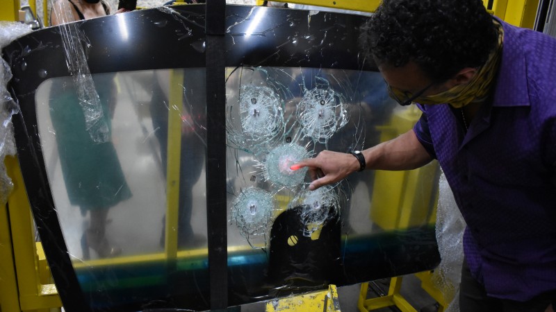 Homem à direita da imagem aponta para marca de disparo central, entre outras quatro marcas, em um vidro de parabrisa de veículo fixado em suporte metálico amarelo.
