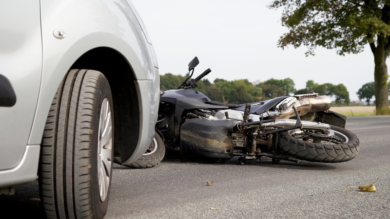 Imagem da lateral de um carro em uma estrada com uma moto caída no chão na frente.