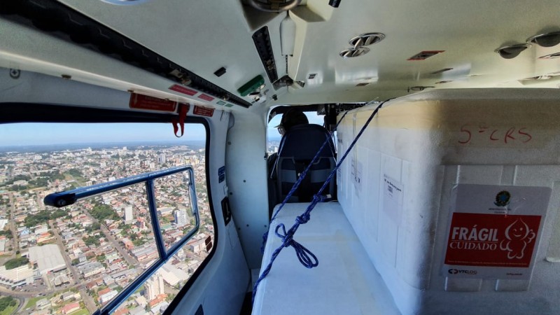 Caixa de isopor com cartaz vermelho com a palavra "frágil" no interior de um helicóptero da BM. Pela janela à esquerda, visão aérea de uma cidade.