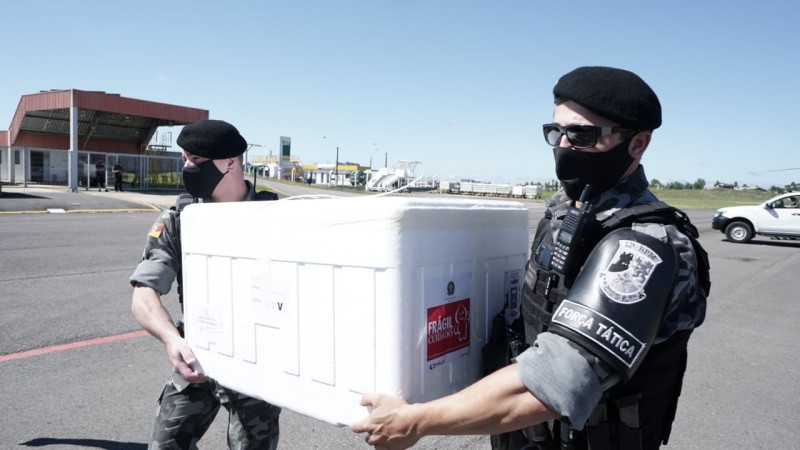 Dois policiais militares carregam uma caixa de isopor com doses da vacina contra Covid-19.