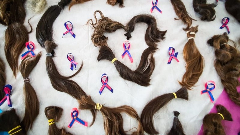 Diversas mechas de cabelo dispostas em uma mesa formando um formato de coração. Ao redor das mechas, há diversas fitinhas das campanhas de conscientização do câncer.