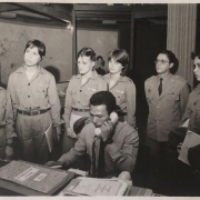 Foto do arquivo pessoal da coronel Cristine. Oficiais em pé, na uma sala, atrás de um servidor que esta ao telefone. 
