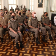 Coronel sentada na sala, onde esta sendo realizada a cerimonia de sua despedida. A sala esta lotada de outros oficiais que a prestigiam neste momento. 