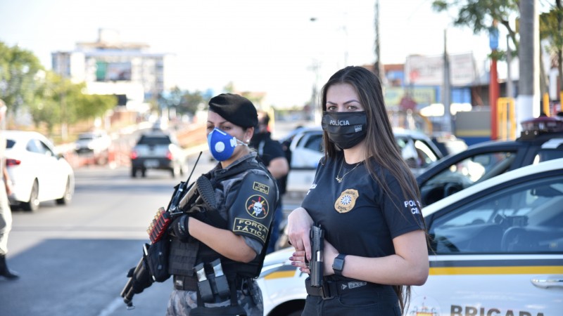 Uma PM e uma policial civil lado a lado, em pé, segurando armas. Ambas usam máscara contra Covid-19. Atrás, viaturas da BM e da Polícia Civil.