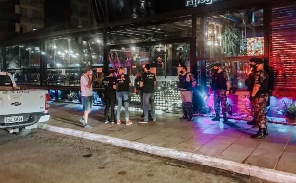 Grupo de policiais civis e militares diante de um restaurante com iluminação cenográfica em via de Capão da Canoa. À esquerda, se vê a traseira de uma caminhonete da Secretaria da Fazenda estacionada junto ao meio-fio.