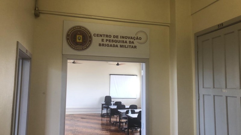 uma porta aberta bem no centro da imagem, dentro estão mesas e cadeiras de uma sala de aula, no umbral uma placa identificando o centro de pesquisa da Brigada Militar