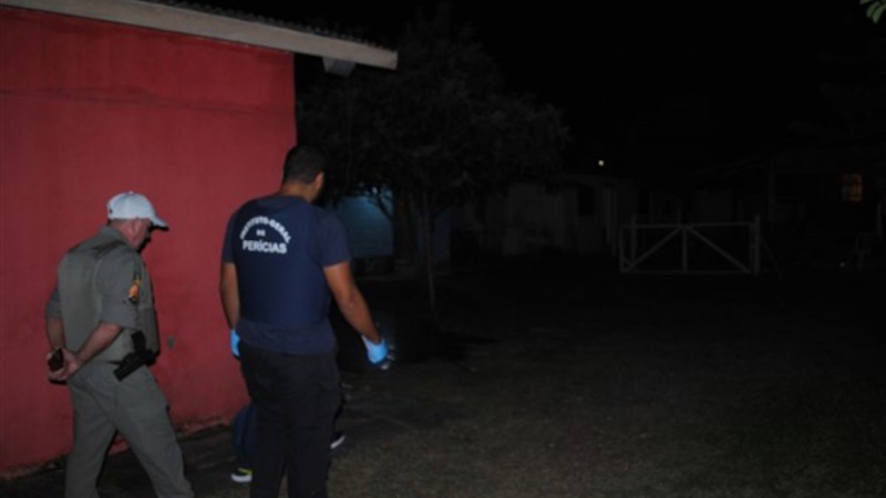 em um ambiente escuro à noite, perito do IGP e policial militar observam cena de crime no pátio de uma residência