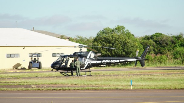Helicóptero da Polícia Civil em um capão de grama. Diante dele, dois homens. Ao fundo, um galpão.