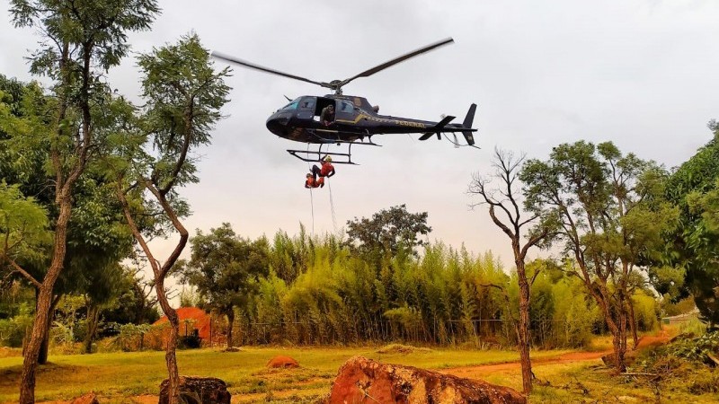 Helicóptero sobrevoando uma área com árvores, com dois bombeiros se segurando na base do helicóptero.