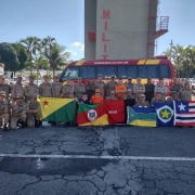 A turma toda dos bombeiros que participaram do curso dispostos em duas fileiras, com alguns de pé e outros abaixados. Os que estão no chão estão segurando bandeiras de alguns estados. Ao fundo, um ônibus dos bombeiros de Goiás.