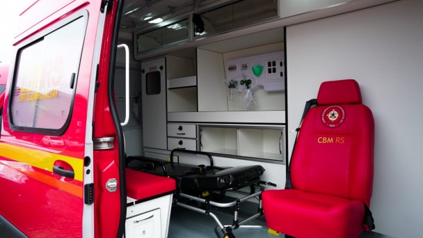 Visualização do interior de uma ambulância de resgate do CBMRS, onde se vê uma cadeira vermelha, uma maca preta e equipamentos de oxigênio em uma bancada branca.