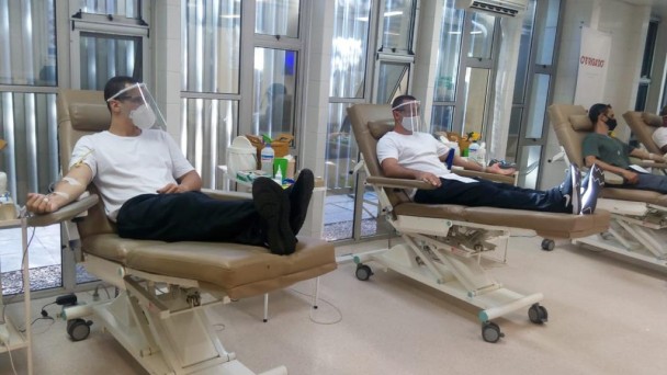 Alunos soldados bombeiros de máscara, face shield, camiseta branca e calça preta sentados em macas hospitalares marrons realizam doação de sangue.