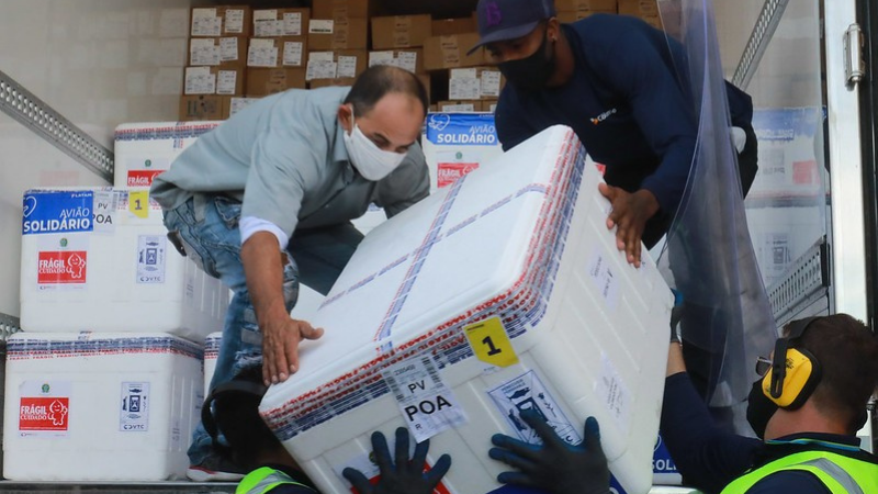 Três homens com máscaras, carregando para um caminhão refrigerado uma caixa com o novo lote de vacinas.