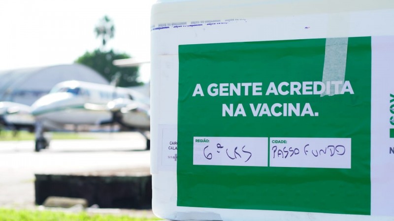 Foto da caixa de imunizantes com um adesivo verde escrito a gente acredita na vacina. Ao fundo o avião da BM