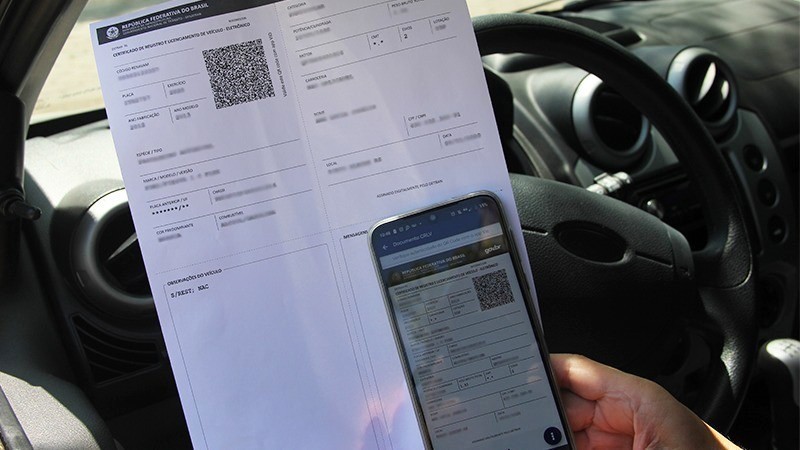 Uma pessoa, sentada dentro de um veículo, no lado do motorista, com um celular e uma folha em mãos, realizando o pagamento da taxa de licenciamento 2021 de forma online.