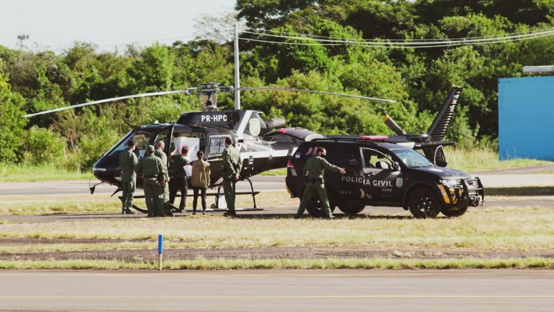 Helicóptero e viatura da Polícia Civil e 7 pessoas auxiliando o carregamento das caixas térmicas.