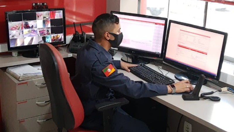Soldado do CBMRS sentado na sala de operções em ferente a dois computadores.