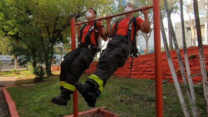 Dois bombeiros militares usando máscara e carregando uma mochila com um cilindro às costas aparecem lado a lado fazendo um exercício de barra fixa no pátio de um quartel do CBMRS.