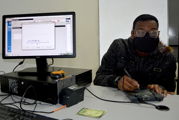 Nicolas Sito, assinando eletronicamente para fazer sua carteira de identidade. Ao seu lado esquerdo, um computador onde aparece sua assinatura e uma máquina para gravar suas digitais.