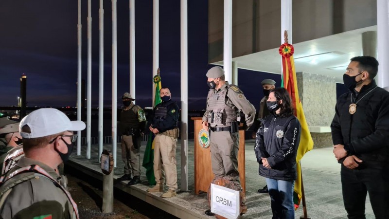 Líderes das instituições envolvidas na operação lado a lado, em pé, diante de um grupo de policiais. Atrás deles, vários mastros de bandeiras.