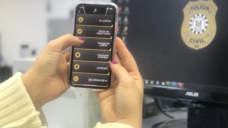 Foto de duas mãos segurando um celular em frente a um desktop, no celular o aplicativo PC Alerta