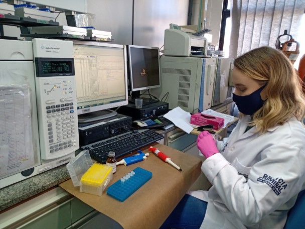 Perita responsável pela análise, Bruna Gauer, aparece de jaleco branco com a marca da perícia criminal, luvas rosas e máscara preta, sentada diante de um computador e outros equipamentos utilizados no trabalho pericial.