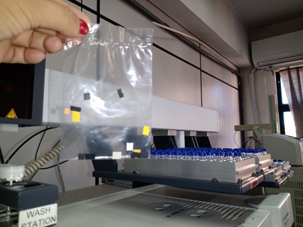 Em primeiro plano, uma mão segura um saco plástico transparente com os selos da droga dentro. Ao fundo, em foco, o cromatógrafo gasoso acoplado à espectometria de massa, que é o equipamento utilizado na análise do material.