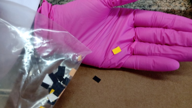 Uma mão com luva rosa segura um selinho amarelo da nova droga. Ao lado, na esquerda, um saco plástico transparente com mais selos amarelos e pretos da droga dentro.