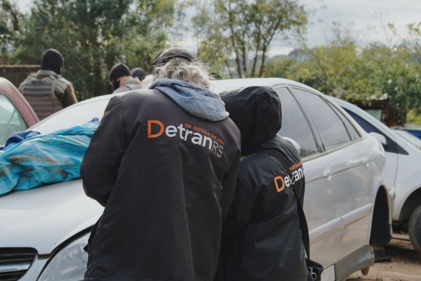 Dois integrantes do DetranRS, de costas, verificando um veículo sedan branco.