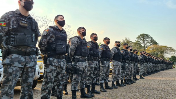 Fila de policiais militares em formação lado a lado no pátio da Academia de Polícia Militar. Todos de máscara.