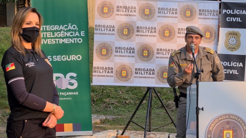 Chefe da Polícia Civil, delegada Nadine Anflor, e comandante-geral da BM, coronel Vanius Cesar Santarosa, diante de banners do RS Seguro, da BM e da Polícia Civil. Santarosa fala ao microfone.