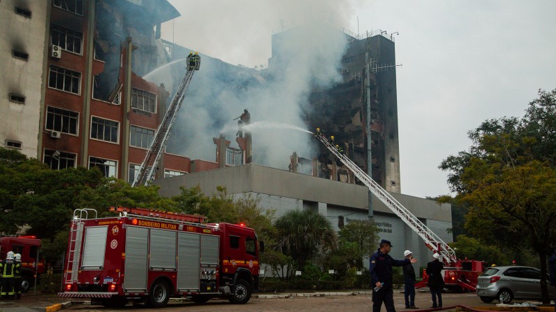 Foto do pr&eacute;dio da SSP no dia 15 de julho, j&aacute; destru&iacute;do pelas chamas. Parte do pr&eacute;dio desabou e os bombeiros ainda trabalham para apagar focos de fogo com dois caminh&otilde;es de combate com longas escadas e mangueiras.