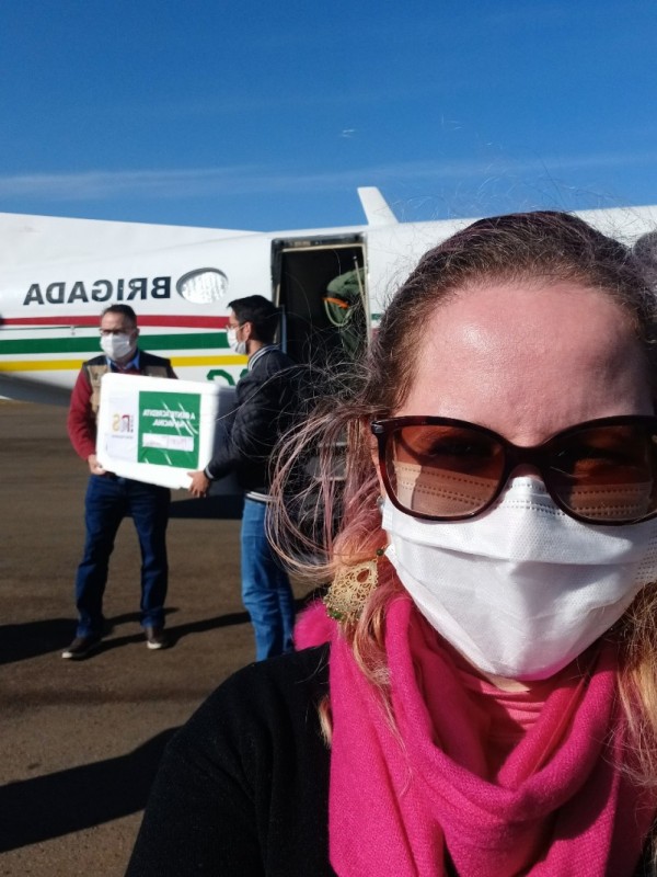 Foto selfie da servidora Marília Bissigo com o avião da Brigada Militar no fundo, de portas abertas, e dois homens carregando uma caixa de isopor com as vacinas.